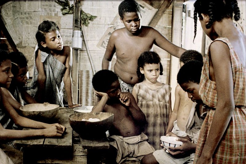 Szenenfoto in Farbe: Eine Gruppe Kinder in einer Hütte stehen und sitzen um einen Tisch mit Essen herum.