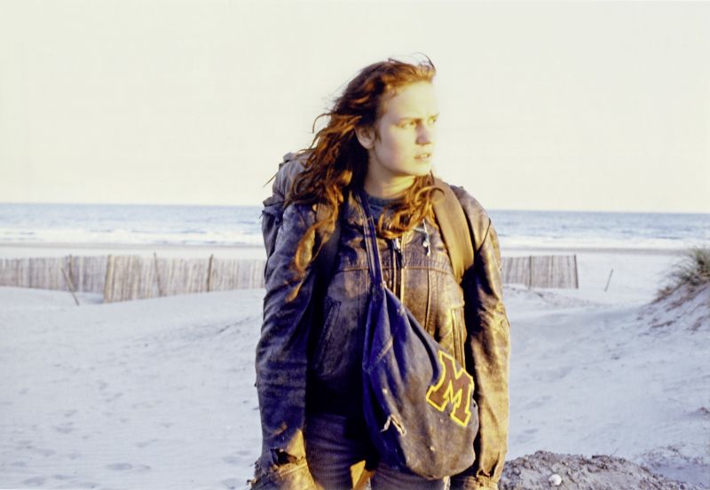 Still in color: A young woman on a beach with a backpack and more luggage, her hair is windswept.
