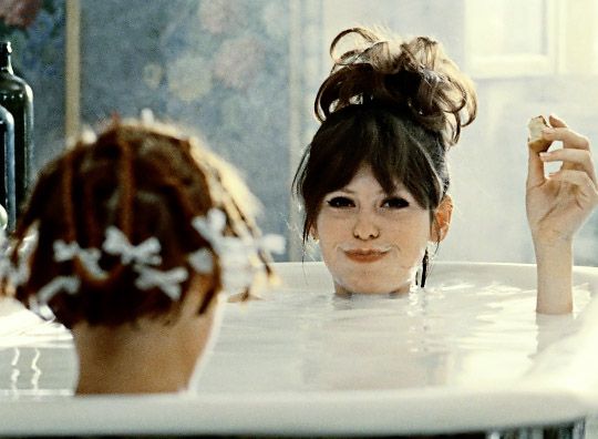 Still in color: A woman sitting opposite another person in a foamie bathtub, only their heads and arms are visible.