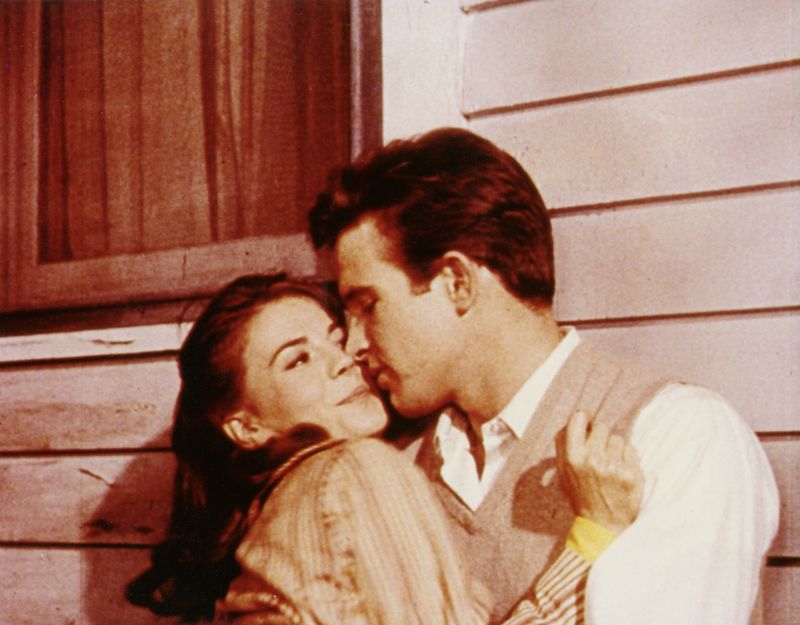 Still in color: A young woman and man, both dark-haired, are standing infront of a wooden house, embracing for a kiss.