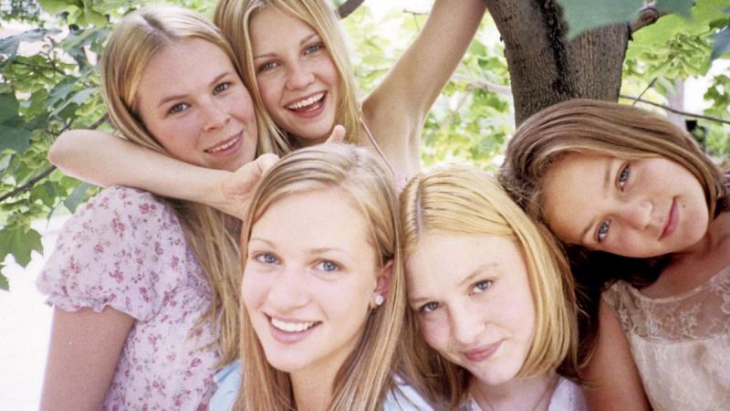 Stil in color: Five blonde girls are standing under a tree, very close togehter and smiling into the camera.