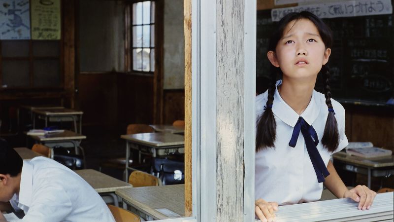Still in color: A young girl in school uniform looking out of the window of a classroom. On the left side behind her another child is sitting at a desk and writing.