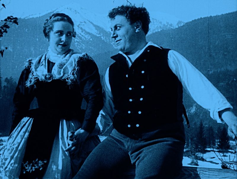 Blau viragiertes (eingefärbtes) Szenenfoto: Eine junge Frau und ein junger Mann, beide in Trachten, im Hintergrund Berge
