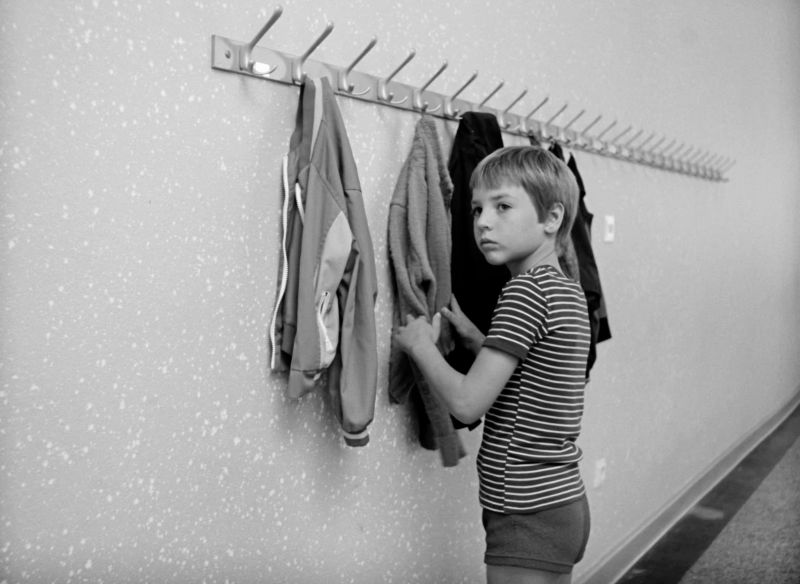 Schwarz-weiß-Szenenfoto: Ein Junge steht an einer Garderobe und fässt eine Jacke an.