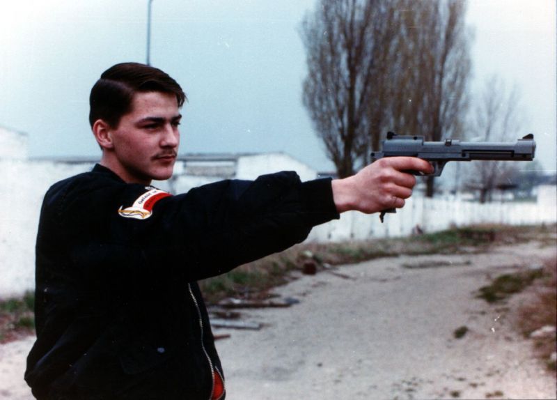 Junge in schwarzer Blousonjacke und Abzeichen des Kaiserreichs hält eine Schusswaffe in der Hand 