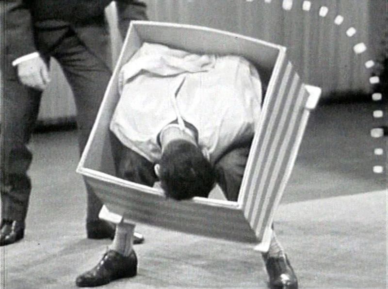 Schwarz-weiß-Foto eines Mannes, vornübergebeugt, der Oberkörper steckt in einem Karton