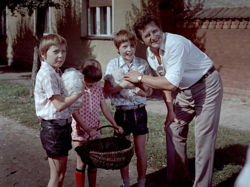 Szenenbild in Farbe: Ein Mann steht mit zwei Jungen, die Hunde auf dem Arm haben, und einem Mädchen, das einen Korb trägt, auf dem Rasen vor einem Haus.