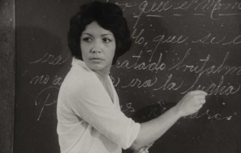Black and white still: A woman is writing on a blackboard while turing back to the room..