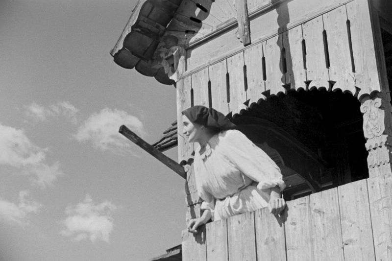Black and white still: A woman in a white dress and a black head scarf is looking through the window of a wooden building into the sky.