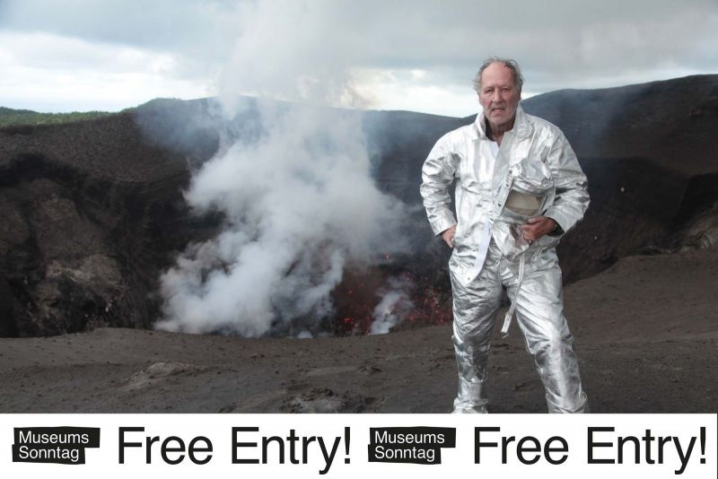 The director stands in protective clothing at the crater of a volcano. Underneath is a banner with the words "museum sunday free entry!"