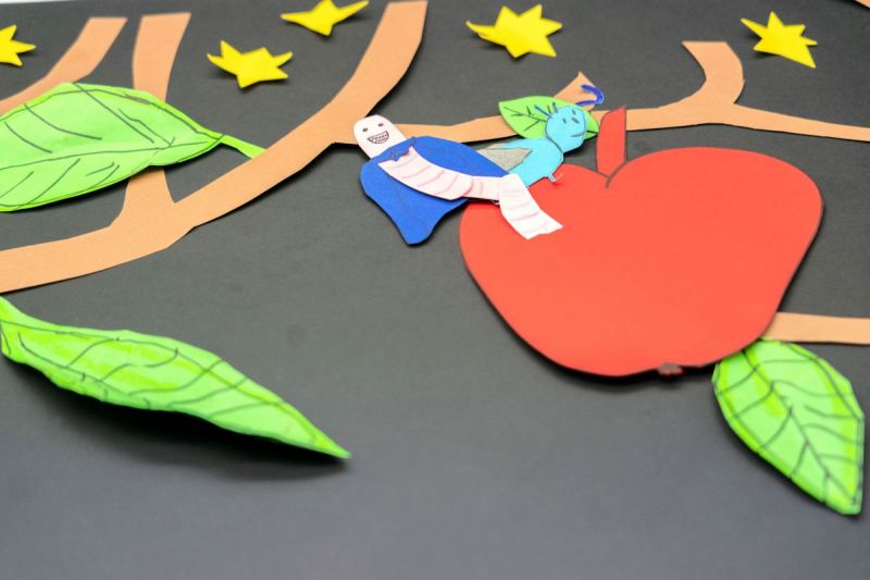 Figures made from colorful paper laid out to form the image of an apple in a tree being eaten by a worm.