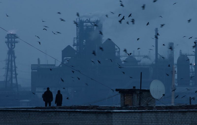 Two people standing on a roof, the silhouette of an industrial plant in the background