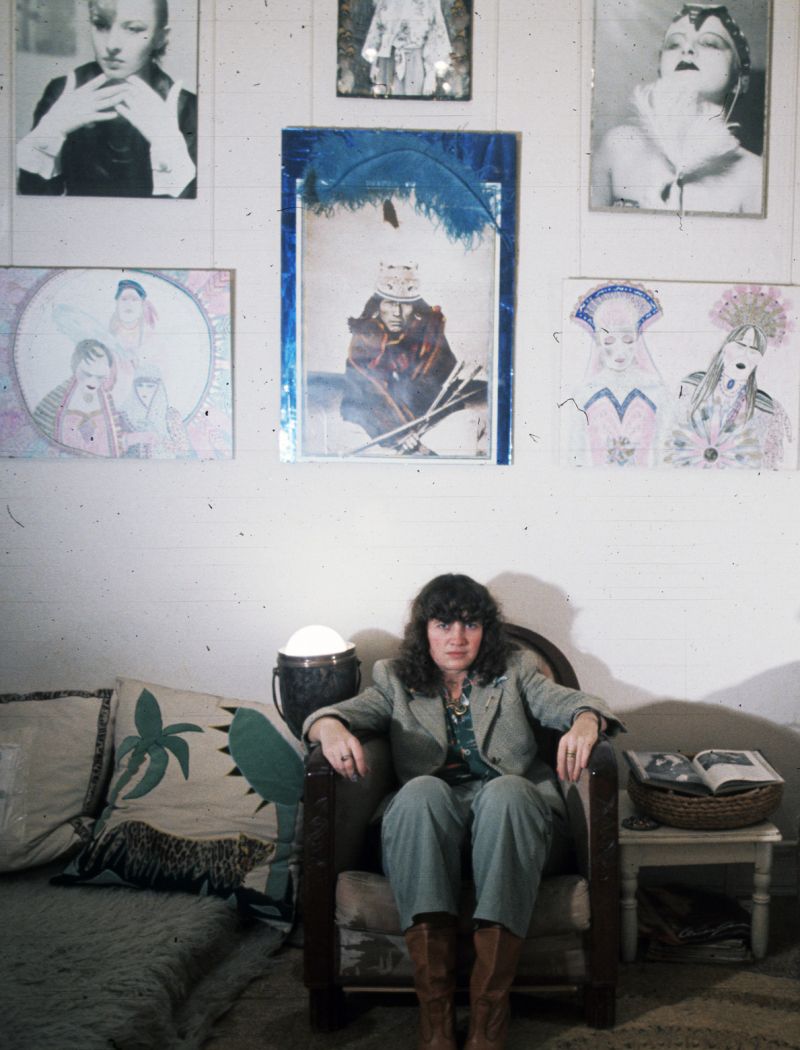 Die Künstlerin sitzt im Sessel, Blick in die Kamera gerichtet. Über ihr an der Wand hängen ihre Kunstwerke.