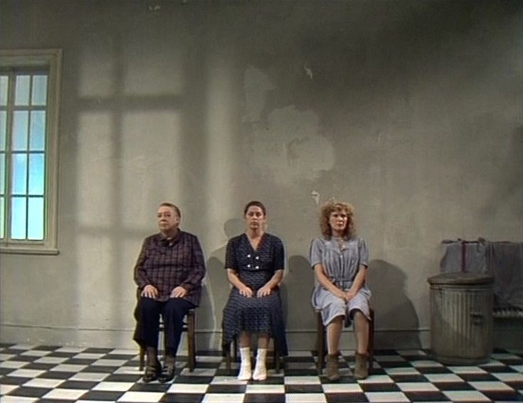 Szenenfoto in Farbe: Drei Frauen sitzen nebeneinander auf Stühlen vor einer grauen Betonwand, der Boden ist schwarz-weiß kariert.