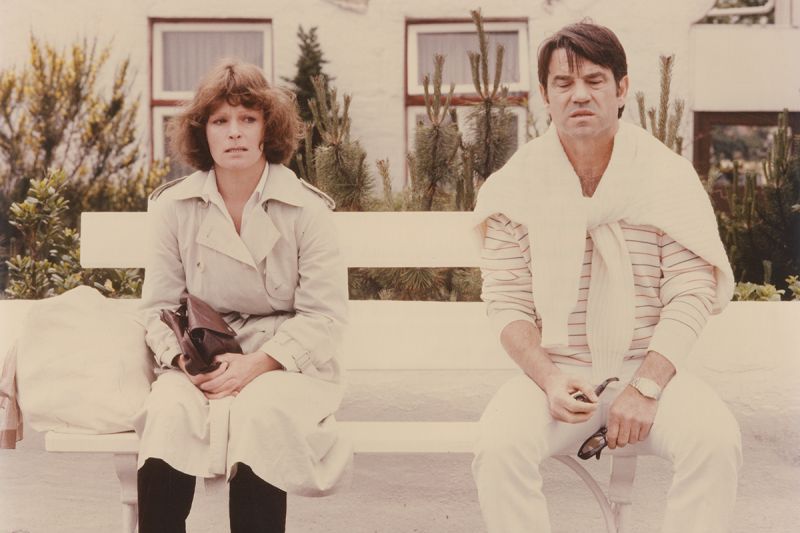 Ein Mann und eine Frau sitzen mit grimmigem Gesichtsausdruck auf einer weißen Bank vor einem Wohnhaus.