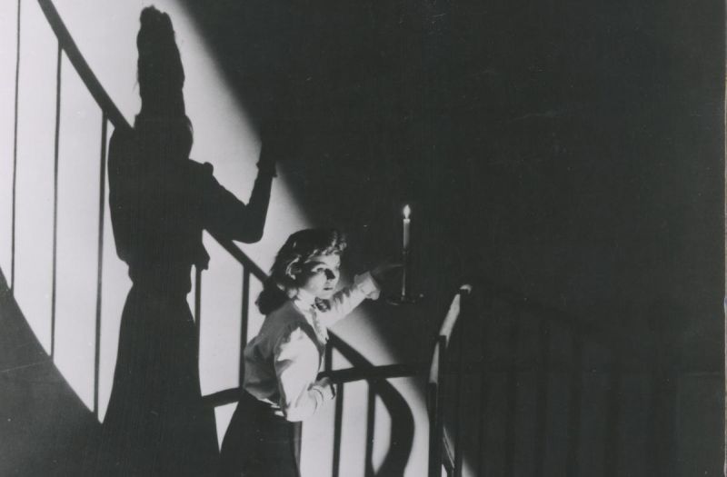Black and white still: A woman, holding a candle, is walking down a spiral staircase, the candlelight is casting a stark shadow on the wall.