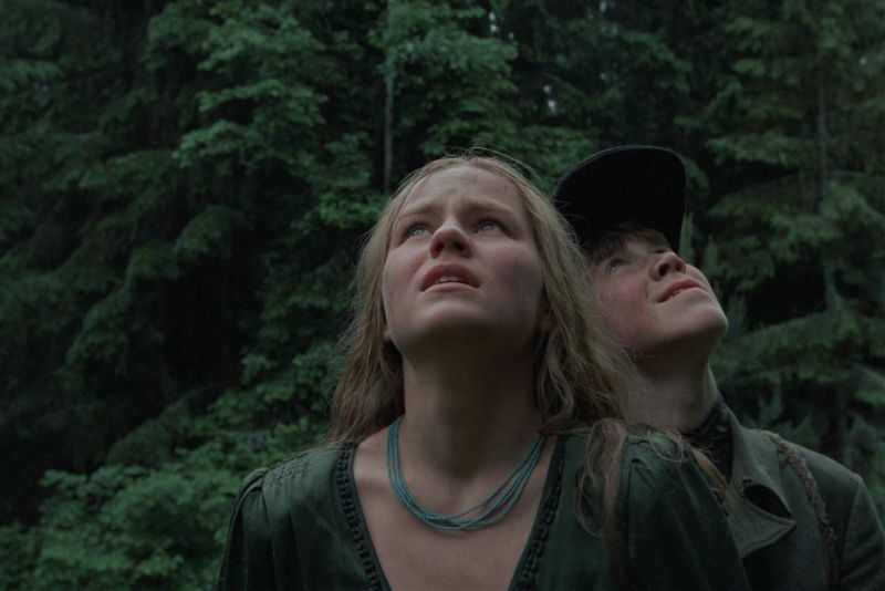 Still in color: A blonde girl and a boy in a dark cap are standing close to each other in a forest and are looking up towards the sky.