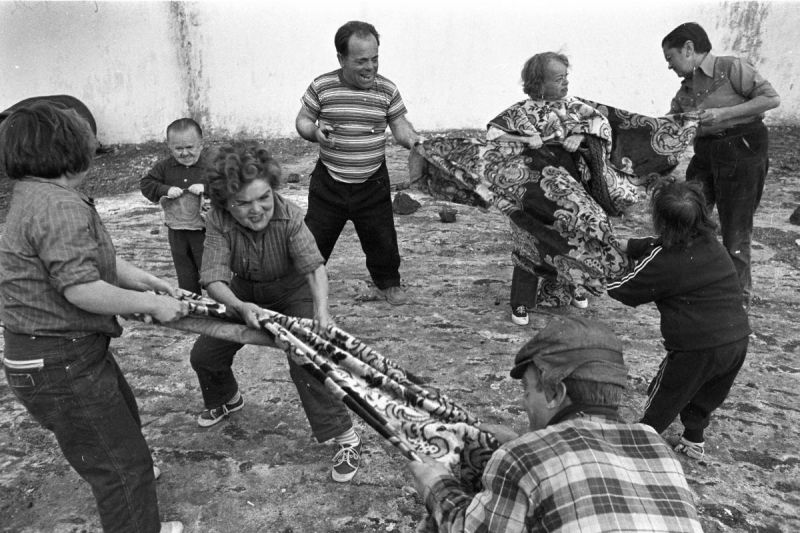 Schwarz-weißes Szenenfoto: Eine Gruppe kleinwüchsiger Menschen spielt draußen mit Tüchern Tauziehen.