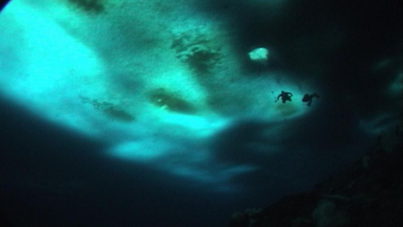 Szenenfoto in Farbe: Eine sehr dunkle Aufnahme unter Wasser von zwei Tauchern, von Links zur Mitte scheint Licht blau durch das Wasser.