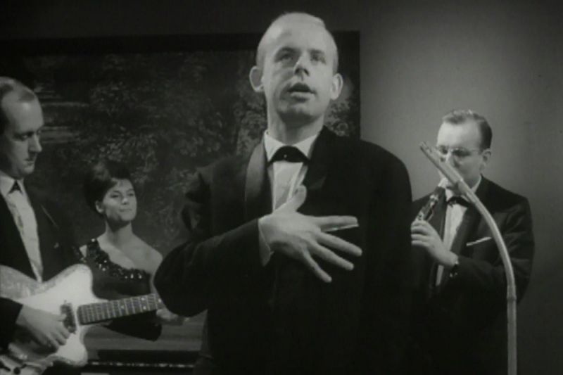 Schwarzweiß-Szenenfoto einer Band bestehend aus drei Männern im Anzug und einer Frau im Kleid