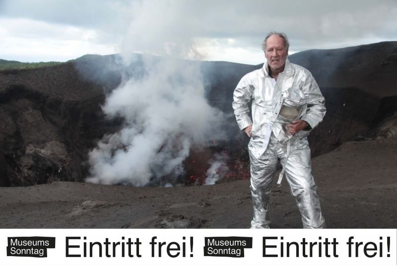 Werkfoto: Der Regisseur steht in Schutzkleidung am Krater eines Vulkans. Darunter ein Banner mit dem Slogan "Museumssonntag Eintritt frei!".
