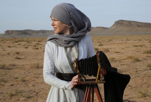 Eine Frau in einem weißen Kleid mit grauem Kopftuch steht in einer Wüste neben einer alten Fotokamera und schaut nach links in die Ferne.