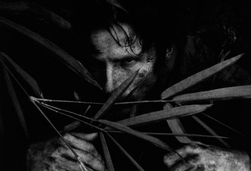 Schwarz-weiß-Foto: Ein Mann guckt grimmig durch die Blätter einer Dschungelpflanze.
