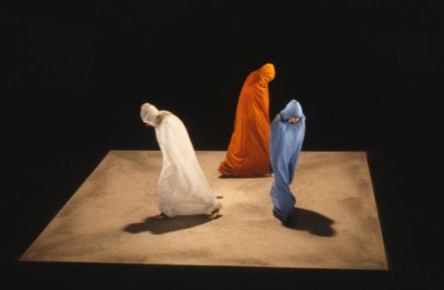 Colour photo: Still from television series Quadrat I, three figures dressed in respectively blue, red and yellow capes walk within a brown square on the ground
