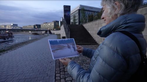 Am Ufer der Spree steht eine Frau und hält ein Foto in die Höhe. Es ist Abend.