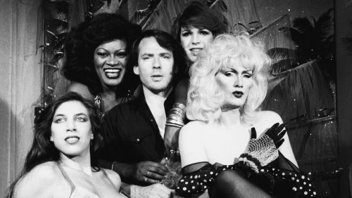 Ein Mann steht inmitten von vier glamourös gekleideten Travestie-Künstlerinnen und trans Frauen