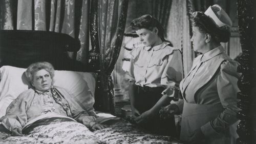 Eine ältere Dame liegt im Bett und sieht voller Wut auf zwei Junge Frauen, die auf der rechten Bildseite an ihrem Bett stehen