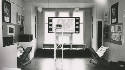 Schwarzweiß-Foto: Ausstellungsraum in Altbau-Wohnraum, am Fenster hängt eine Leinwand, auf die ein Filmbild projiziert wird.
