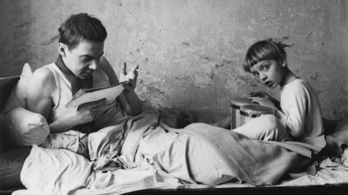 Ein Mann und ein Junge auf dem Bett. Der Mann liest ein Buch und raucht eine Zigarette, der Junge spielt Bongos.