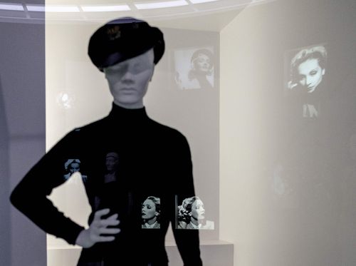 Puppe mit schwarzem Marlene-Dietrich-Kostüm hinter Glas, in dem sich Porträtfotos der Schauspielerinen spiegeln