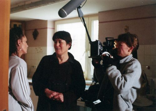 Three women standing in a bathroom, two in conversation while a third is filming them.