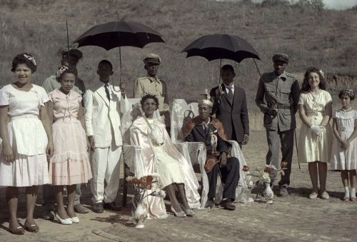 A group of people are standing around two others, who are sitting on chairs, and holding two dark umbrellas.