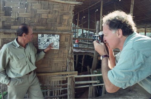 Werner Herzog is taking a photo of Dieter Denger who is pointing at a photograph pinned to a wooden wall.