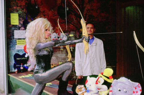 A blonde woman wearing a tight bodysuit and corsage is standing shooting gallery holding a bow and arrow.