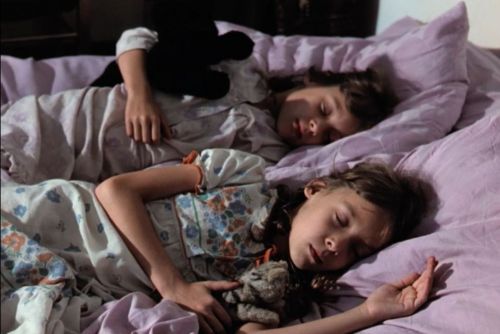 Still: two young girls sitting lying next to each other in bed, asleep. One is holding a toy, the sheets are light purple.