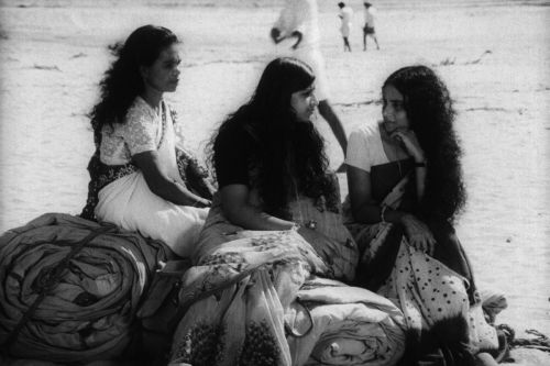 Black and white still: Three women dressed in saris are sitting on rolled up blankets on the floor