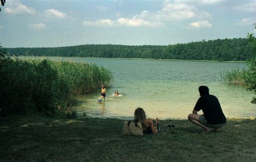 A man at woman are sitting at the shore of a lake and watching two children play in the water.