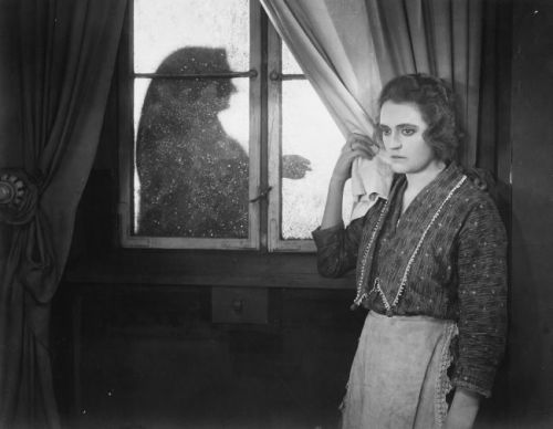 Schwarz-weißes Szenenfoto: Eine traurig blickende Frau steht an einem Fenster und hält den Vorhang zurück, auf der anderen Seite erkennt man eine weitere Person.