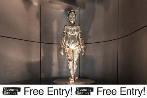View of the permanent exhibition: Silver manneqiun of the "Maschinen Maria" from the film 'Metropolis' with the banner "Museum Sunday Free Entry!" underneath..