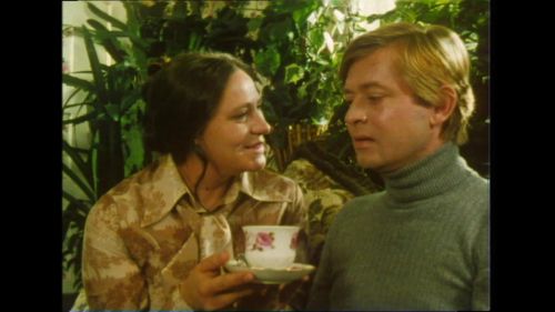 Szenenfoto in Farbe: Vor einer großen Zimmerpflanze sitzen ein Mann und eine Frau nebeneinander, die Frau hält eine Tasse in der Hand als ob sie sie dem Mann geben will.