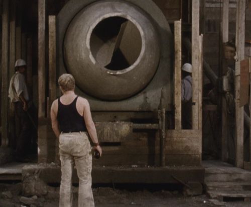 Still in color: A man is facing away from the camera towards a large cement mixer, there are two other men standing to the right and left.
