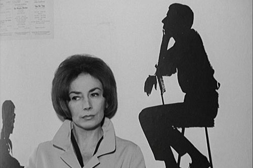 Black and white still: portrait of a woman in front of a white wall, on the right side behind here, there is the shadow of a man sitting on a chair.