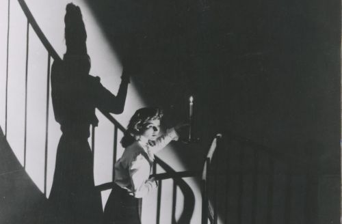 Schwarz-weißes Szenenfoto: Eine Frau läuft mit einer Kerze in der Hand eine Wendeltreppe hinab. Das Kernzenlicht wirft einen starken Schatten der Situation an die Wand.