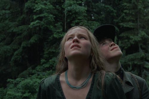 Szenenfoto in Farbe: Ein blondes Mädchen und ein Junge mit dunkler Kappe stehen im Wald dicht nebeneinander und blicken nach oben.
