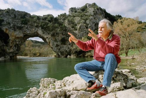 Still in color: A man with grey hair and a mustache sitting on a rock in front of a lake, gesturing with his hands while talking.