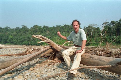 Still in color: The director sitting on a fallen tree in a cleared area in front of a forest.